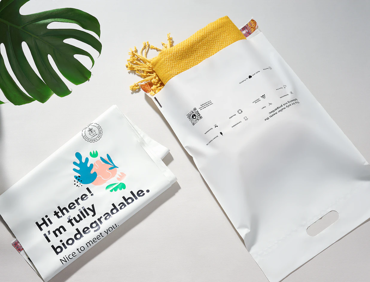 Flat lay of white bio poly mailers printed with eco properties and 'Hi there! I'm fully biodegradable.', one holding a yellow blanket next to a monstera leaf
