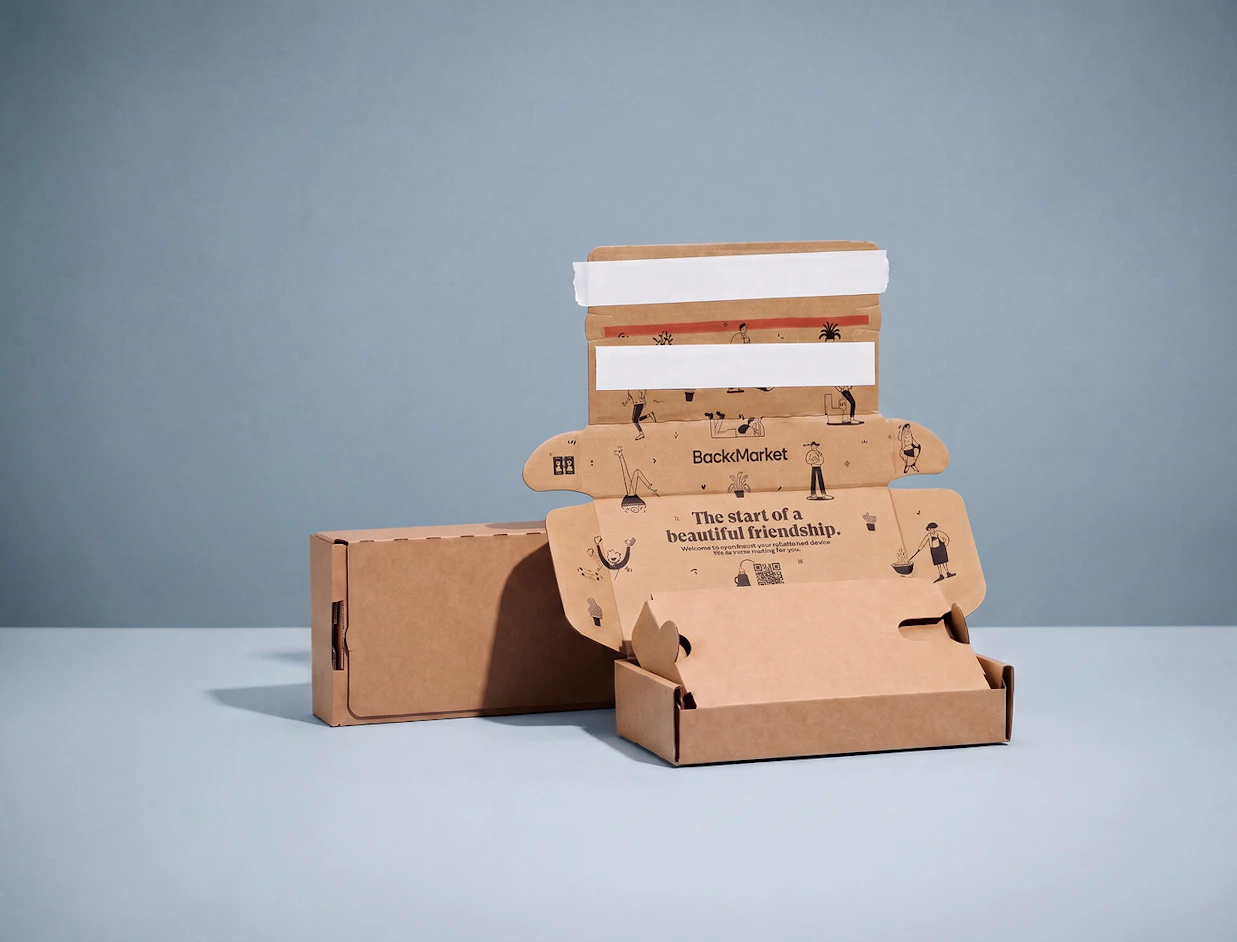 An open brown cardboard phone packaging box branded BackMarket with inner print 'The start of a beautiful friendship.' next to a closed box on a light blue background
