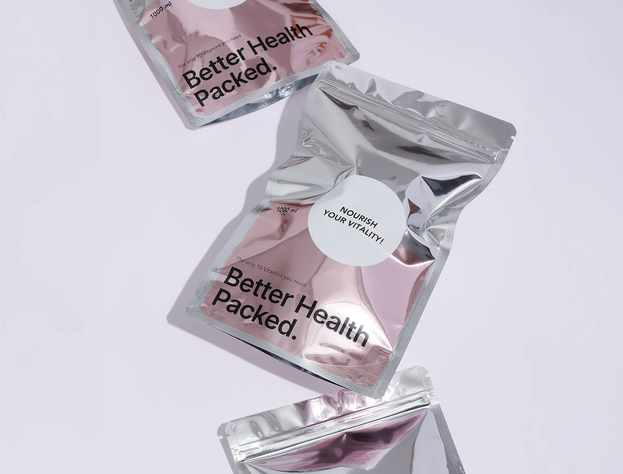 Shiny metallic silver foil ziplock pouches printed with 'Better Health Packed.' and round white sticker labels on a light purple surface