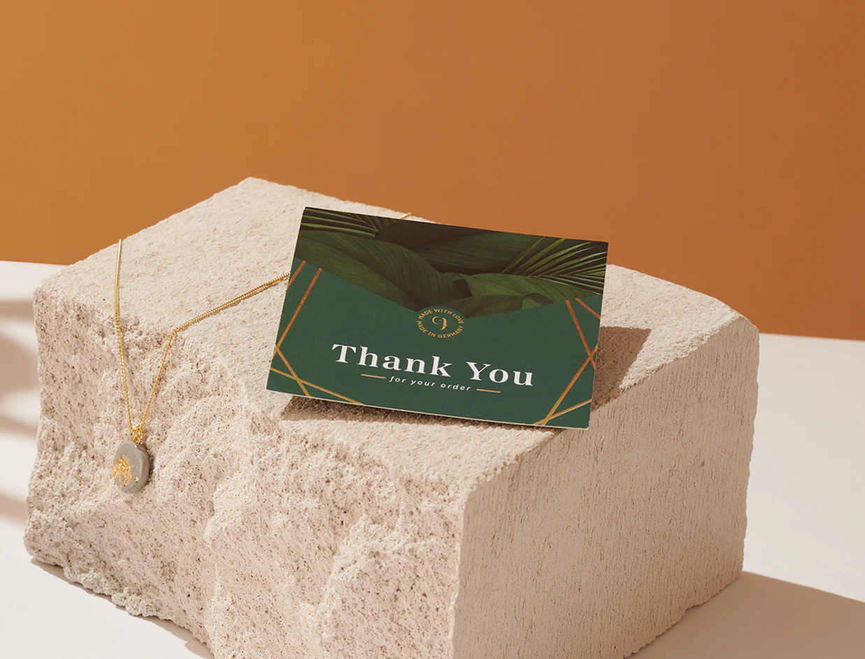 A green 'Thank You for your order' card resting on a stone display block next to a gold pendant necklace
