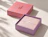 An open two-piece gift box with a pink grid lid and a purple base containing tissue paper sealed with a 'nest' sticker