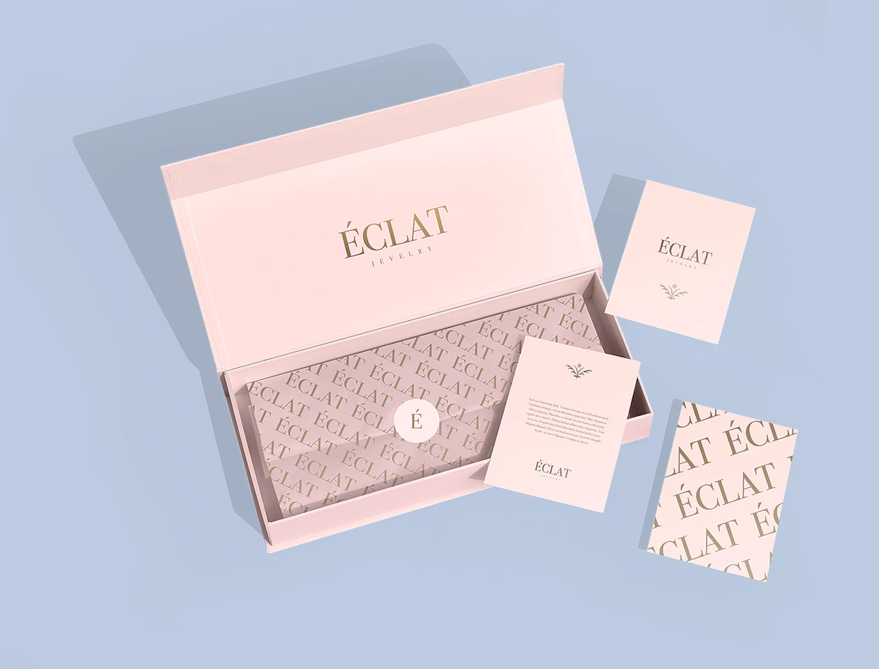 An open light pink magnetic closure box branded 'ÉCLAT JEWELRY' with printed tissue paper, custom cards, and a sticker seal on a blue background