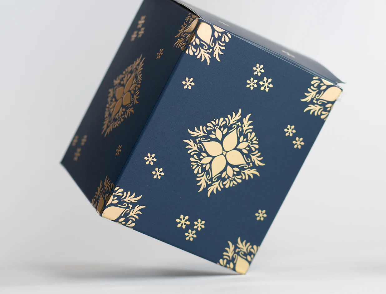 Dark blue cube product box decorated with intricate gold floral and snowflake patterns, tilted against a white background