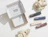 An open white mailer box branded 'XLASH COSMETICS' surrounded by rocks and four boxed beauty products on a light background