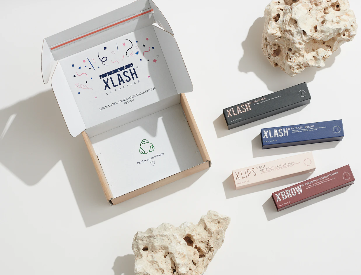 An open white mailer box branded 'XLASH COSMETICS' surrounded by rocks and four boxed beauty products on a light background