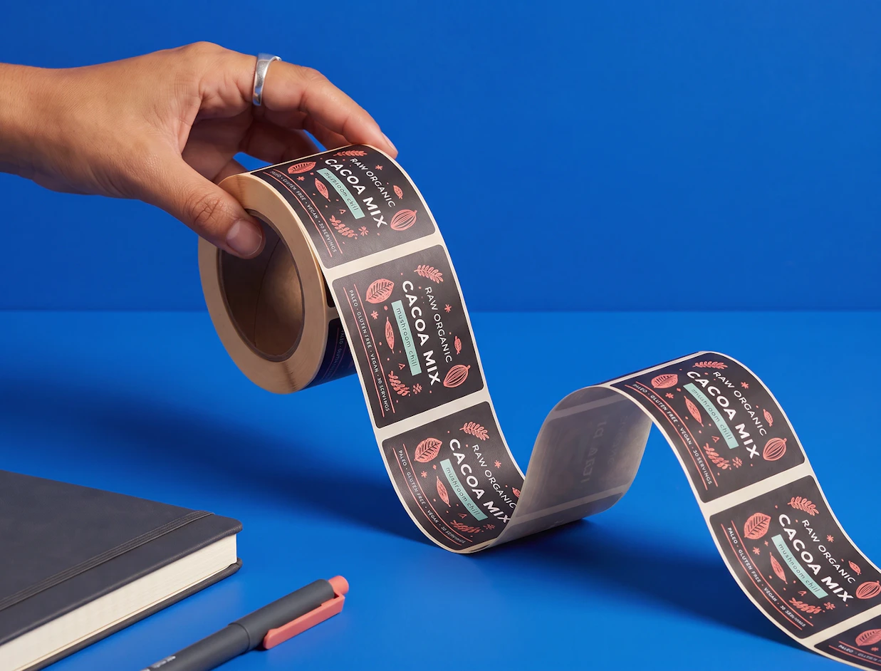 A hand holding an unrolled roll of dark brown product labels that read 'RAW ORGANIC CACOA MIX'