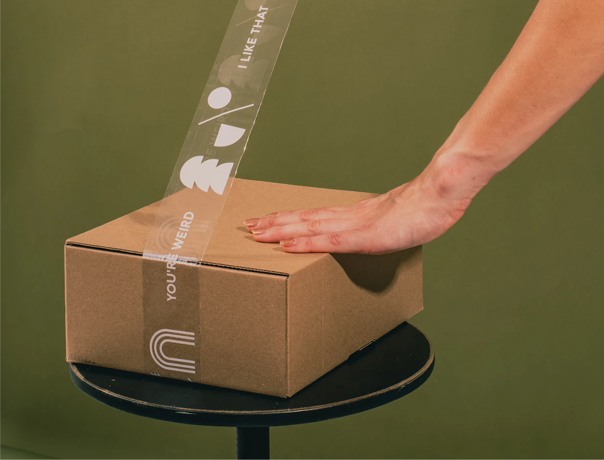 A hand smoothing transparent tape with white text 'YOU'RE WEIRD I LIKE THAT' onto a cardboard box on a stool