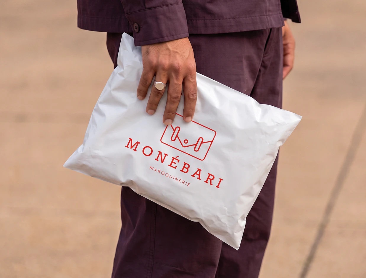 A person in a dark outfit holding a white poly mailer bag printed with red 'MONÉBARI MAROQUINERIE' branding