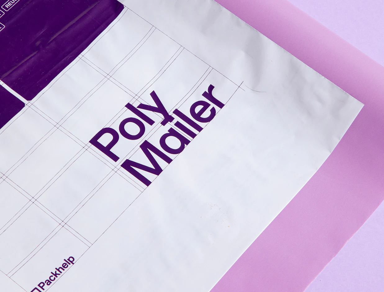 Close-up of a white poly mailer with purple grid lines and 'Poly Mailer' typography, branded with Packhelp on a pink background