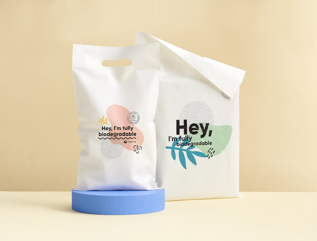 Two white biodegradable poly mailers printed with colorful abstract shapes and 'Hey, I'm fully biodegradable' text displayed on a blue cylindrical pedestal