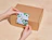 Two hands applying a patterned green and blue sticker with the word 'OAKEN' onto a cardboard shipping box