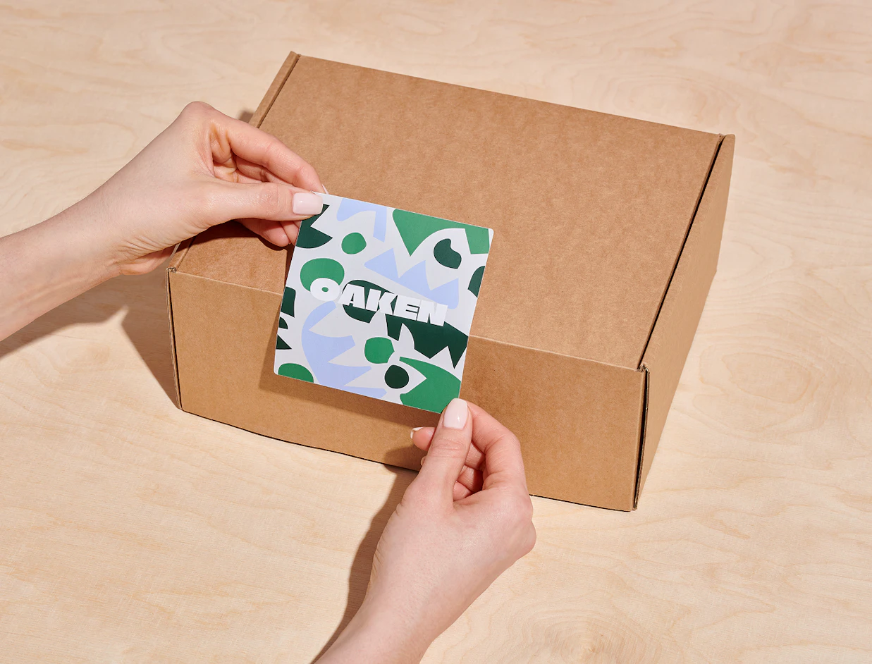 Two hands applying a patterned green and blue sticker with the word 'OAKEN' onto a cardboard shipping box