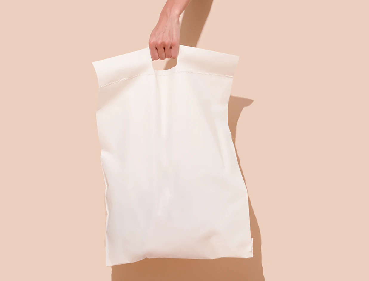 A hand holding a plain white padded poly mailer bag with a die-cut handle against a beige background