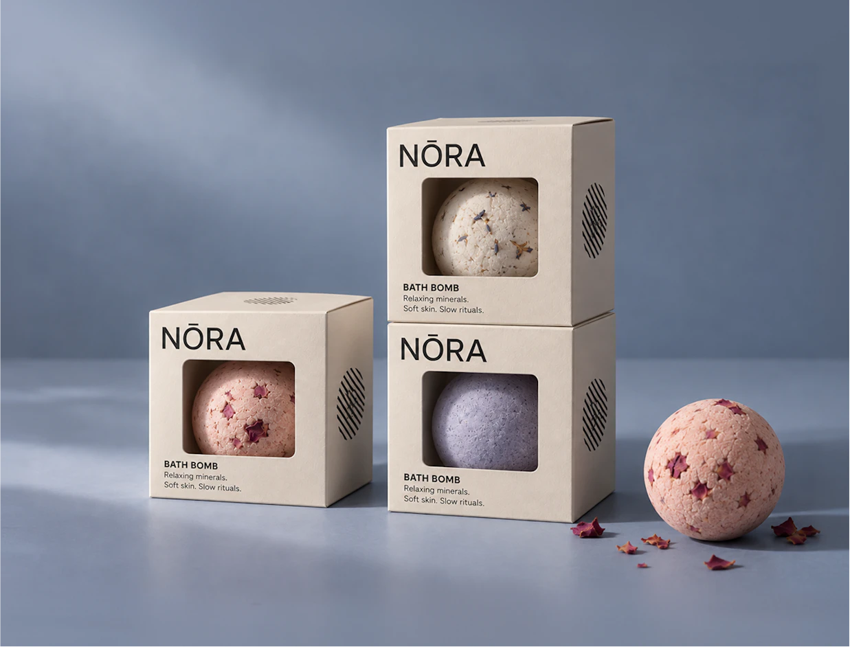 Three beige NŌRA bath bomb boxes with cutout windows displaying textured bath bombs inside, set against a muted blue backdrop