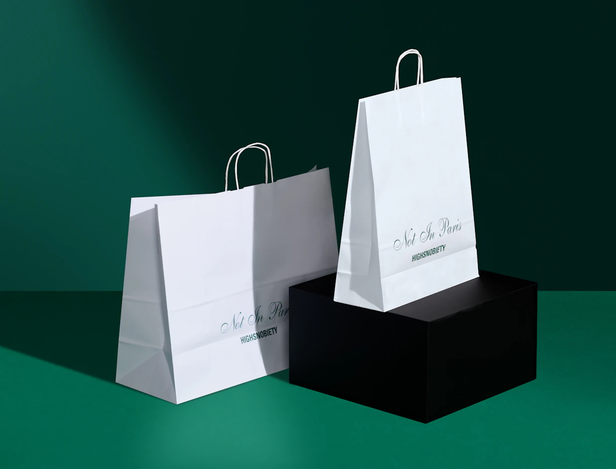 Two white printed paper bags with twisted handles and a green print on a dark green backdrop