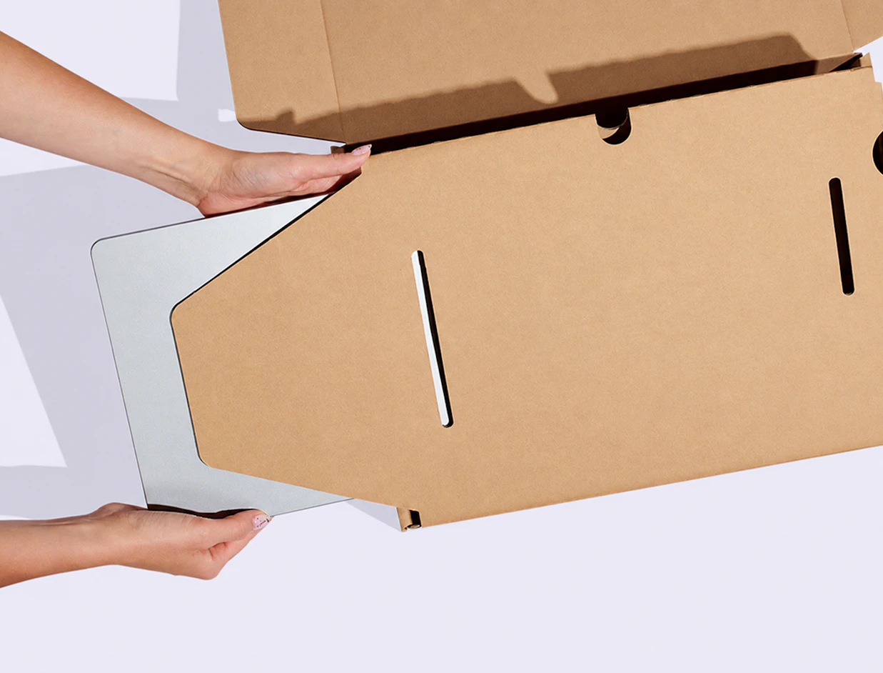 Top-down view of hands placing a laptop into a protective kraft cardboard insert inside a mailer box on a white background