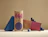 Two kraft paper pillow boxes printed with colorful geometric graphics and social media handles, displayed with blue geometric props on a beige surface