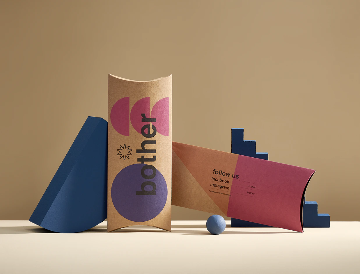Two kraft paper pillow boxes printed with colorful geometric graphics and social media handles, displayed with blue geometric props on a beige surface