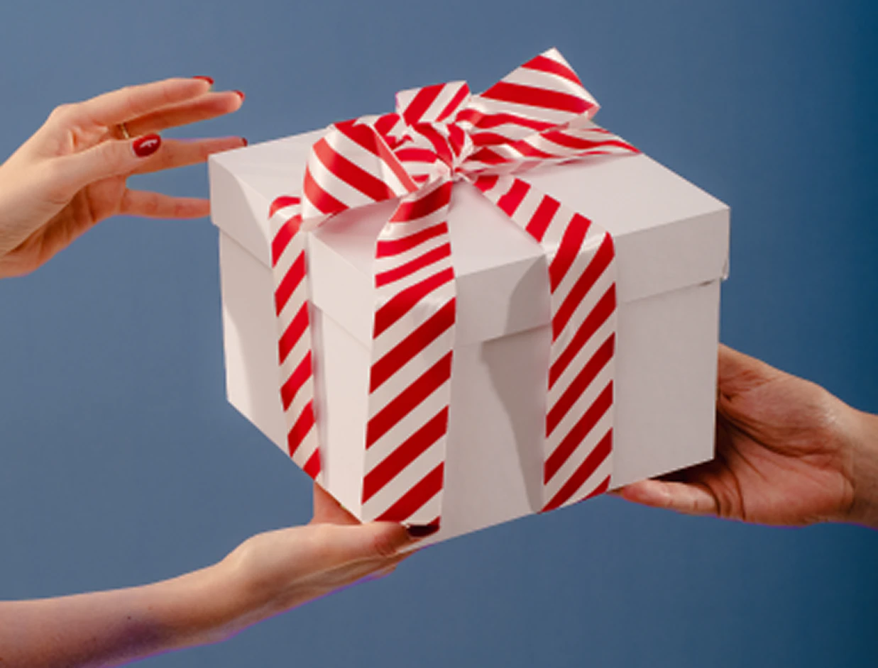 Hands handing over a white gift box wrapped with a red and white diagonal striped ribbon bow on a blue backdrop