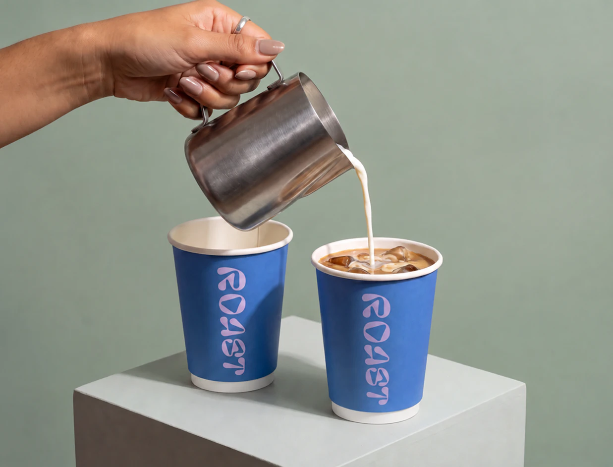 A hand pouring milk from a metal pitcher into a blue paper cup labeled 'ROAST' filled with iced coffee